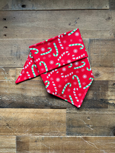 A red bandana with a green and white candy cane and snowflake pattern, folded on a rustic wooden plank background.
