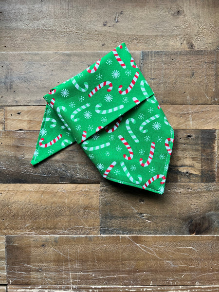 A green fabric bandana with a festive holiday print of candy canes, snowflakes, laid flat on a rustic wooden plank surface.