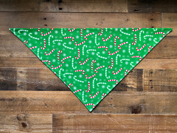 A green triangular pet bandana with a festive pattern of red and white candy canes and white snowflakes, laid flat on a rustic brown wooden plank background.