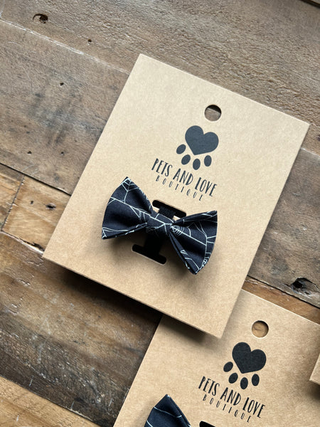 A black and white Spider web patterned pet bow tie displayed on a brown kraft paper card with the brand logo 'PETS AND LOVE BOUTIQUE', set on a rustic wooden surface.