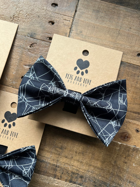A black bow tie with a Spider Web pattern, displayed on a brown kraft paper card printed with the brand 'Pets and Love Boutique' and a paw print logo, set on a rustic wooden surface.