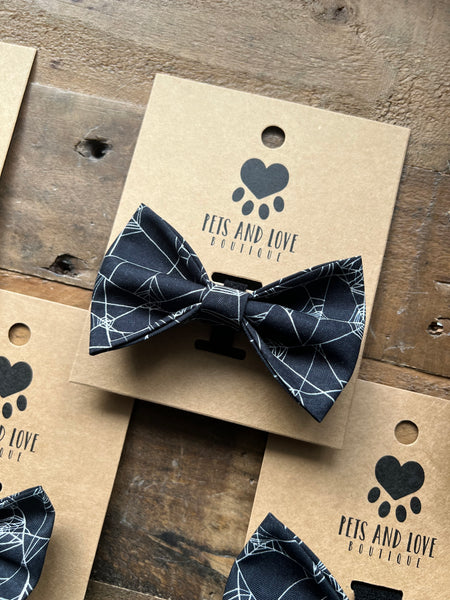 A black pet bow tie with a white Spider Web pattern, displayed on a brown kraft paper card printed with the brand logo 'Pets and Love Boutique'.