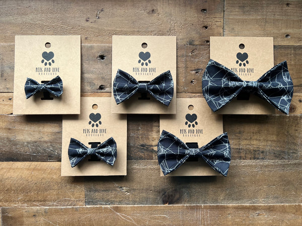 Five black and white spider web patterned bow ties on brown kraft paper cards, arranged on a rustic wooden background. 