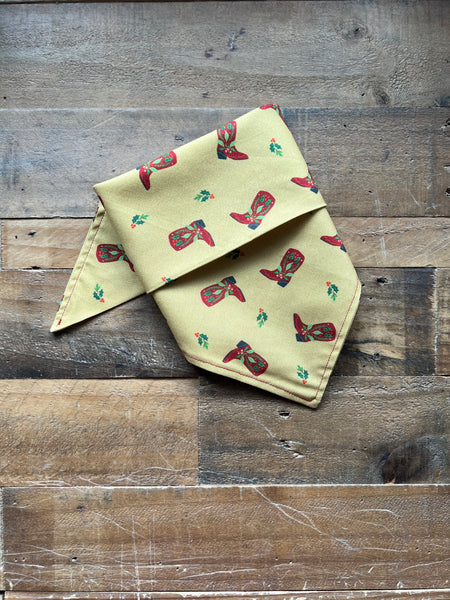 A folded gold bandana featuring a repeating pattern of red cowboy boots and small green holly leaves, displayed on a rustic wooden plank surface.