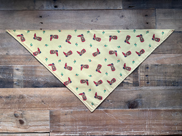 A triangular bandana gold fabric with a repeating print of red cowboy boots and small green leaves, laid flat on a rustic wooden plank background.