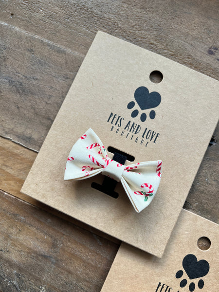 A small pet bow tie with a cream base and red and green holiday candy cane print, displayed on a brown kraft paper card printed with 'Pets and Love Boutique' and a black paw print logo, set on a rustic wooden surface.