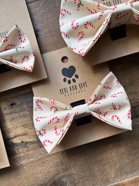 Three cream-colored pet bowties with a red and green candy cane pattern, displayed on brown kraft paper cards on a rustic wooden surface. The center card features the black logo and text 'PETS AND LOVE BOUTIQUE'.