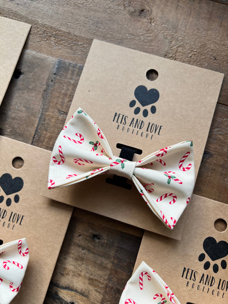 A cream pet bow tie with a red and green candy cane and holly pattern, displayed on a brown kraft paper card printed with the brand 'PETS AND LOVE BOUTIQUE'.