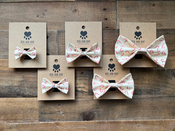Five dog bow ties with a Candy Cane print on a cream fabric, displayed on brown kraft paper cards with the brand logo 'Pets and Love'. The cards are arranged on a rustic wooden plank background
