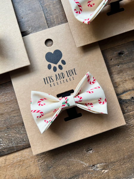 A cream pet bow tie with a red and green candy cane and holly pattern, displayed on a brown kraft paper card with the brand 'Pets and Love Boutique' printed on it, set on a rustic wooden surface.