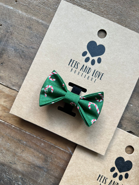 A green fabric pet bow tie with a candy cane pattern, attached to a brown kraft paper display card printed with the brand name 'Pets and Love Boutique'.