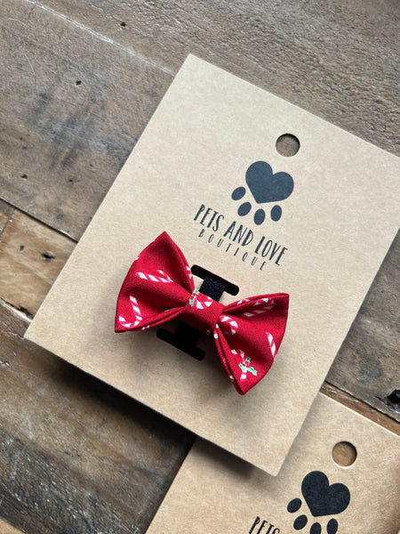 A red pet bow tie with a candy canes pattern, attached to a brown kraft paper display card printed with the brand name 'Pets and Love Boutique'. The card is placed on a rustic wooden surface.