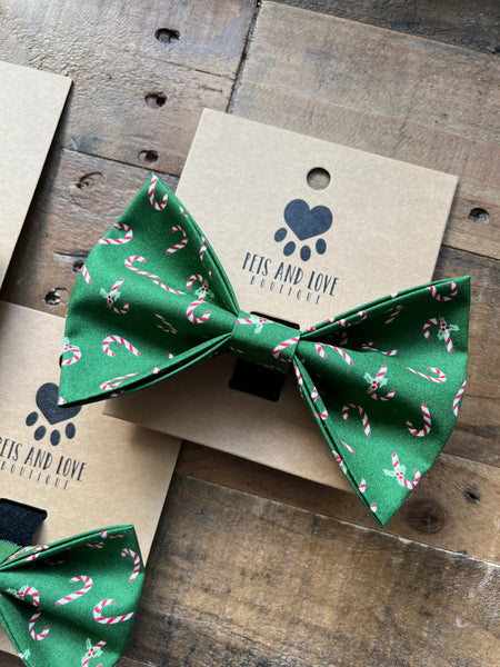 A green pet bow tie with a candy cane pattern, displayed on its kraft paper card which features the 'PETS AND LOVE  logo. The bow tie is photographed on a rustic wooden surface.