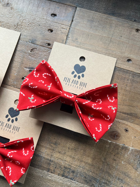 A red pet bow tie with a white candy cane pattern, displayed on a kraft paper card labeled 'PETS AND LOVE ACCESSORIES' on a rustic wooden surface.