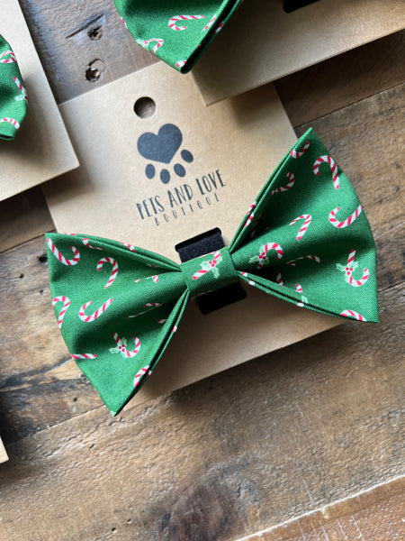A green pet bow tie with a candy cane pattern, displayed on a kraft paper card printed with the brand name 'PETS AND LOVE' on a rustic wooden surface.
