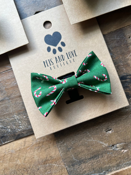 A green pet bow tie with a candy cane pattern, displayed on a kraft paper card from 'Pets and Love Boutique' on a rustic wooden surface.