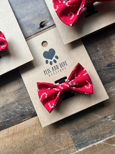 A red candy cane patterned pet bow tie displayed on a kraft paper tag with the brand name 'PETS AND LOVE ACCESSORIES' printed on it, arranged on a rustic wooden surface.