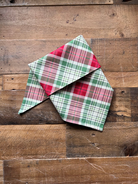 A folded plaid bandana in red, green, and cream, displayed on a rustic wooden surface.