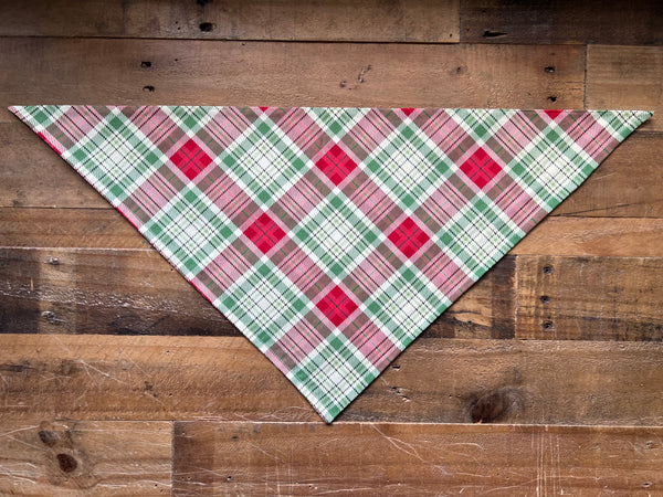 A triangular pet bandana with a festive red, green, and cream plaid pattern, laid flat on a rustic wooden plank background.