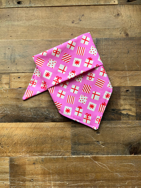 A pink pet bandana with a white and red gift box pattern, laid flat on a rustic wooden plank surface.