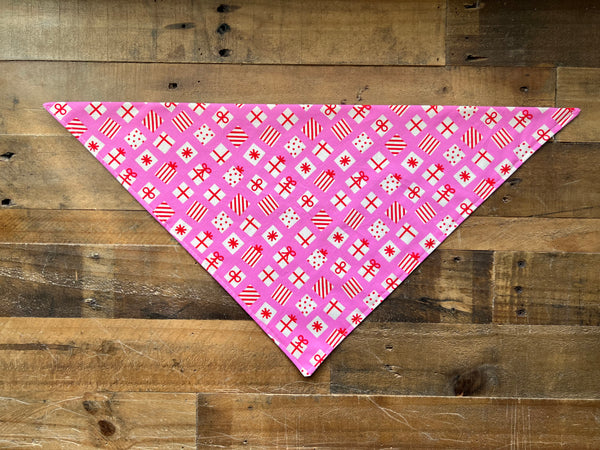 A pink triangular bandana with a festive red and white holiday pattern, laid flat on a rustic wooden plank background.