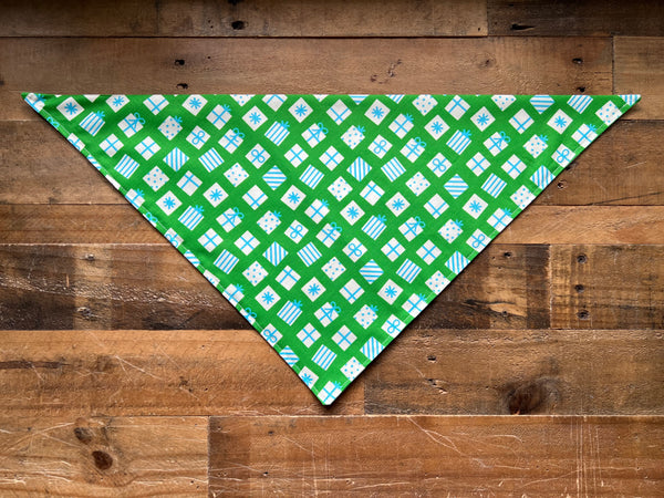 A green triangular bandana with a repeating pattern of white and light blue gift boxes, laid flat on a rustic brown wooden plank background.