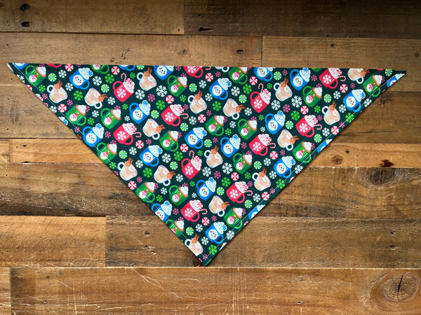 A triangular pet bandana with a colorful holiday pattern of mugs, snowflakes, and Candy Canes on a dark background, laid flat on a rustic wooden plank surface.