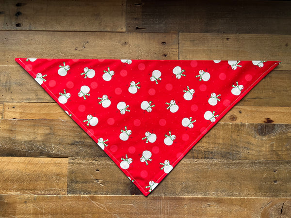A red triangular pet bandana with a white snowman pattern, laid flat on a rustic wooden plank background.
