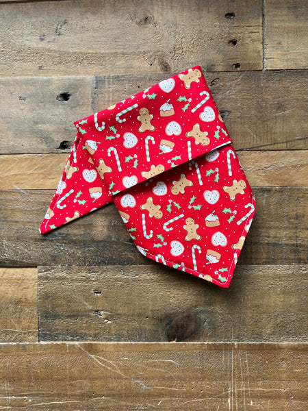 A red pet bandana with a festive gingerbread and candy cane print, laid flat on a rustic wooden plank background.