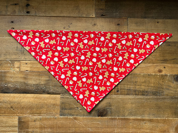 A red triangular pet bandana with a festive holiday print of gingerbread men, candy canes, and snowballs, laid flat on a rustic wooden plank background.