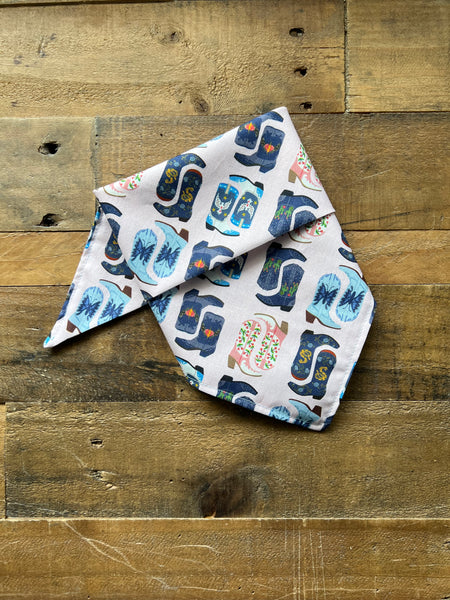 A folded white bandana or scarf with a colorful pattern of blue and navy boots, pink flowers, and brown details, displayed on a rustic wooden plank surface.