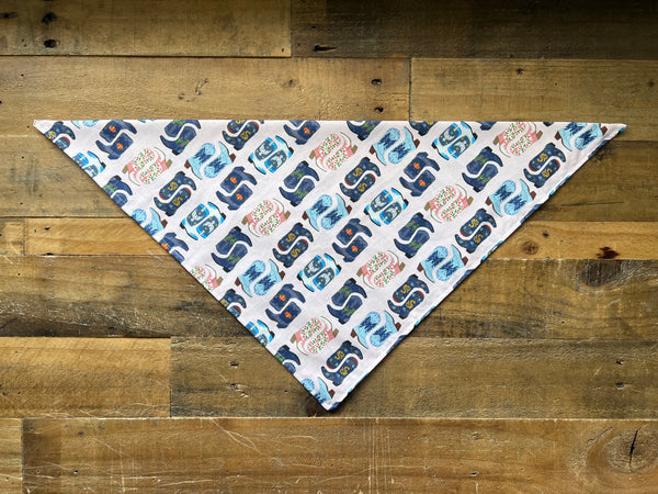 A triangular pet bandana with pink background and a repeating pattern of blue, navy, pink boots laid flat on a rustic wooden plank surface.