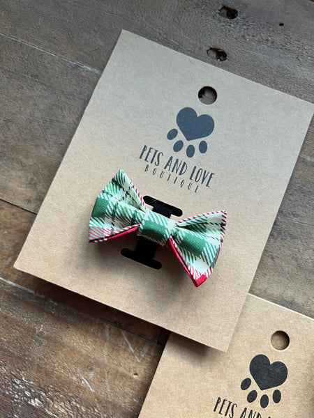 A small pet bow tie with a green, cream, and red plaid pattern, attached to a brown kraft paper display card printed with the brand name 'PETS AND LOVE BOUTIQUE'. The card is placed on a rustic wooden surface.
