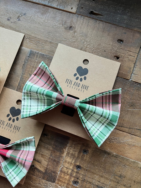 A plaid pet bow tie in green, red, and cream, tied on a brown kraft paper display card with the brand name 'Pets and Love Boutique' printed on it, set on a rustic wooden plank surface.