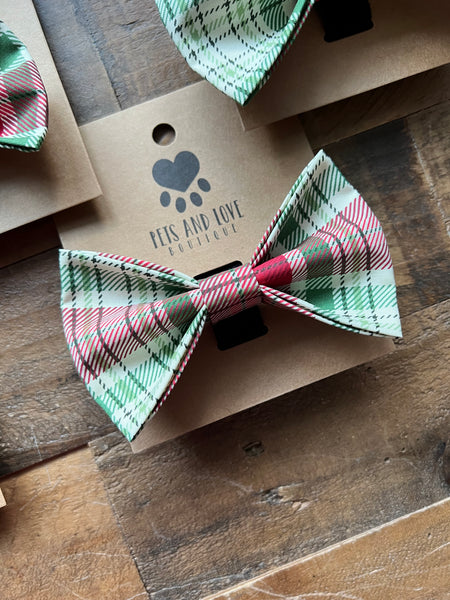 A plaid pet bow tie in green, red, and cream, displayed on a brown kraft paper card with the brand name 'PETS AND LOVE BOUTIQUE' printed on it, set on a rustic wooden surface.