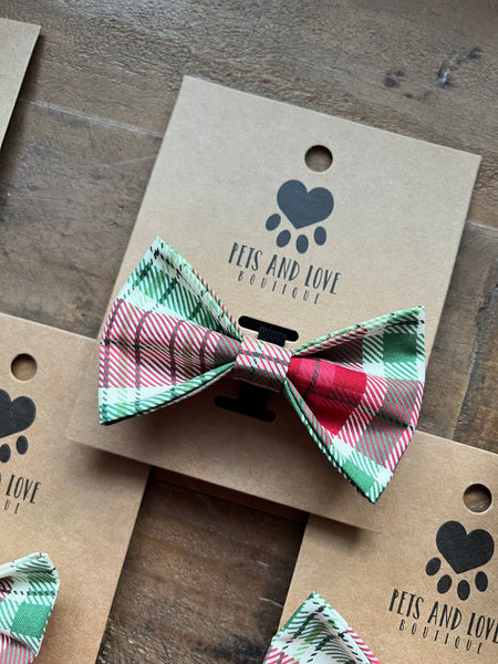 A plaid pet bow tie in red, green, and cream, attached to a brown kraft paper display card with the brand name 'PETS AND LOVE BOUTIQUE' printed in black. The card is placed on a rustic wooden surface.