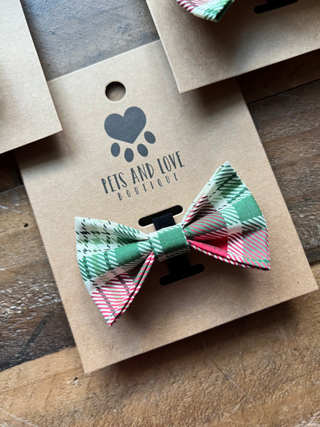 A pet bow tie with a green, red, and cream plaid pattern, displayed on a brown kraft paper card printed with the brand name 'Pets and Love Boutique' on a rustic wooden surface.