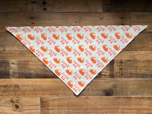A triangular bandana beige fabric with a repeating pattern of orange 'CUTIE PIE' text and pie illustrations, laid flat on a rustic wooden plank background.