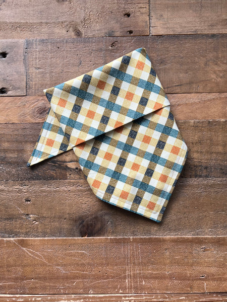 A folded bandana with a blue, orange, and cream gingham plaid pattern, displayed on a rustic wooden surface.