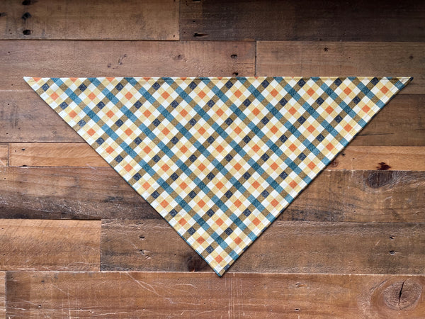 A triangular dog bandana with a blue, orange, and cream plaid pattern, laid flat on a rustic wooden plank background.