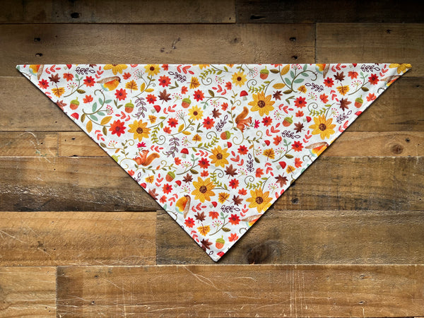 A triangular bandana with a colorful autumn floral and bird pattern, laid flat on a rustic wooden plank background.