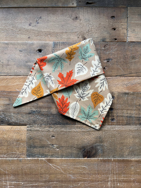 A folded fabric bandana or pet bandana with an autumn leaf pattern in orange, teal, yellow, and beige, brown background, displayed on a rustic wooden plank surface.