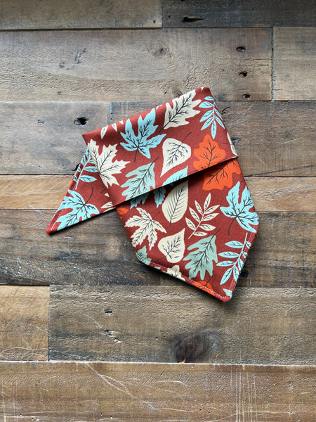 A folded fabric bandana or pet bandana with an autumn leaf pattern in rust red, light blue, and cream, displayed on a rustic wooden plank background.