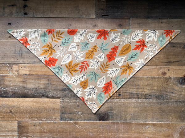 A triangular bandana-style pet accessory with an all-over autumn leaf pattern in orange, teal, mustard, and beige on a brown background, laid flat on a rustic wooden plank surface.