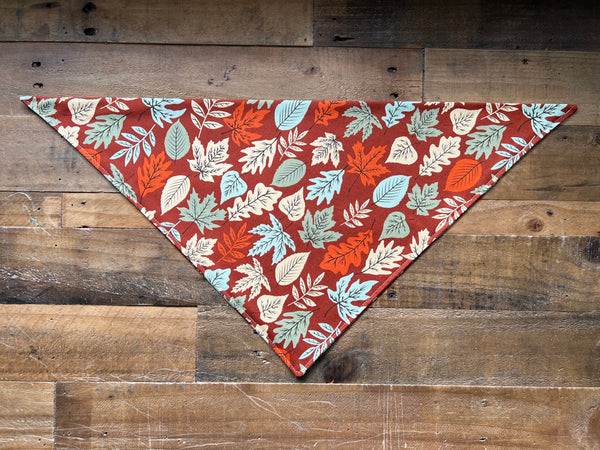 A triangular pet bandana with an autumn leaf pattern in rust red, orange, and light blue, laid flat on a rustic wooden plank background.