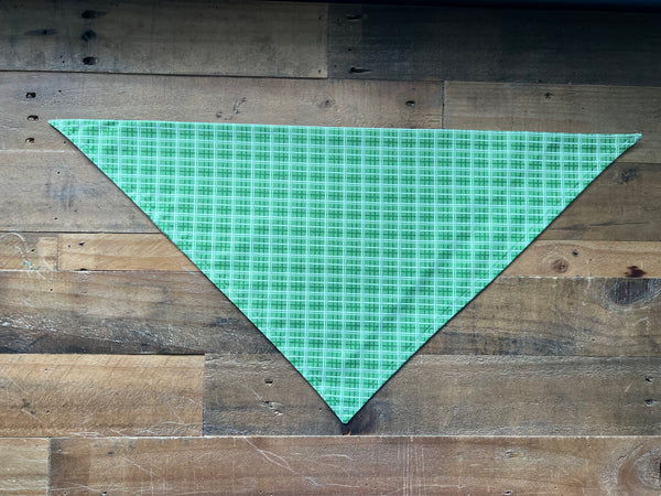 A green and white plaid triangular bandana, likely for a pet or scarf, laid flat on a rustic wooden plank background.