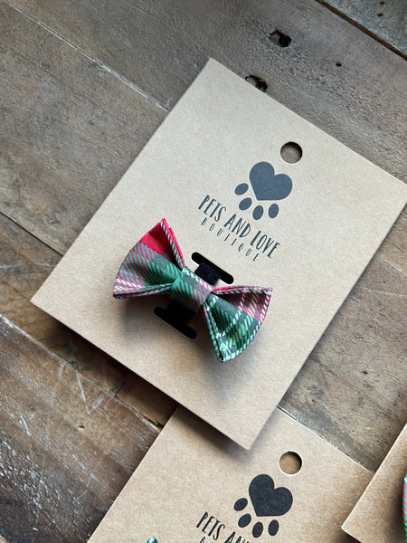 A small plaid bow tie for pets, featuring a green, red, and white tartan pattern, displayed on a brown kraft paper card with the brand name 'PETS AND LOVE BOUTIQUE' printed on it, set on a rustic wooden background.