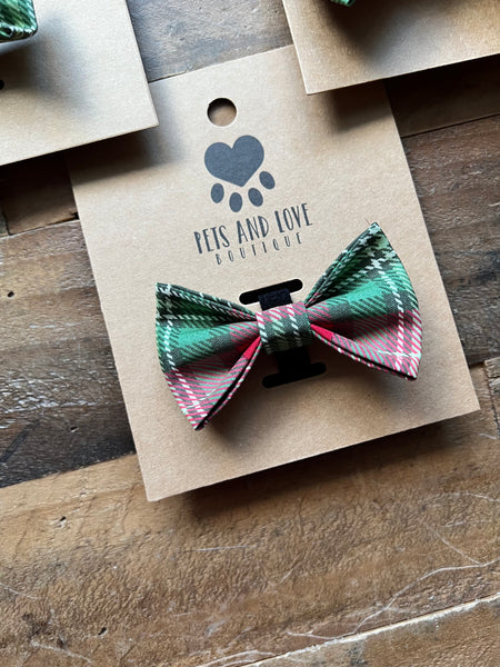 A pet bow tie in a green, red, and white plaid pattern, attached to a brown kraft paper display card printed with the brand name 'Pets and Love Boutique' and a paw print logo, set on a rustic wooden surface.