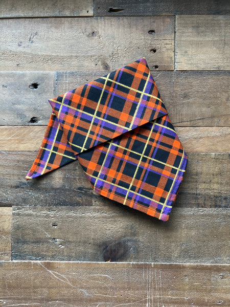 A plaid bandana in orange, black, purple, and gold, folded and displayed on a rustic wooden plank background.