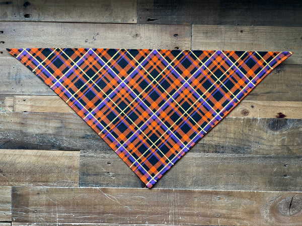 A triangular pet bandana with a vibrant orange, purple, gold and black plaid pattern, laid flat on a rustic wooden plank background.
